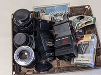 Overview of lot items inside woven basket, including several camera lenses, two camera flashes, lens caps, Black's waterproof film, and camera accessory packaging.