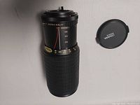 Osawa zoom lens 75-260mm with front lens cap beside it on white background showing side profile and zoom markings.