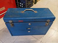Blue metal toolbox closed, showing handle and clasp latches with some wear and rust spots.