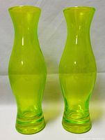 Pair of two translucent yellow-green Vaseline glass vases standing side by side on a white textured background, showing front profile.