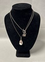 Front view of two silver necklaces displayed on black velvet stand, showing beaded choker with toggle clasp and clear teardrop crystal pendant on chain.