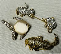 Photo showing three brooches: rhinestone cat and mouse costume brooch, cat's eye fish brooch, and filigree fish brooch