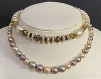 Two vintage 18 inch beaded costume necklaces displayed on a white jewelry bust. The top necklace features cream pearls with gold-toned textured beads and decorative dark silver and black beads. The bottom necklace features round faux pearls in cream, lavender, and silver.