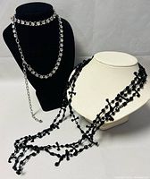 Two costume necklaces displayed on bust stands: silver tone chain with clear crystals and multi-strand black beaded necklace.