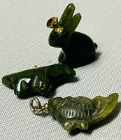 Three small carved jade pendants shaped as animals laid on a textured surface, showing their detailed carving and metal loops.