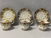 Three teacups with matching saucers arranged in a row, displaying distinct floral patterns and gold trim.