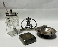 Overview of four antique silver plated items on neutral background, including a crystal jam jar with silver lid and spoon, a silver plated napkin holder with windmill motif, a small footed bowl, and a square trinket box.