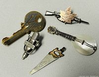 Five vintage costume jewelry pieces including key, microphone, saw, string instrument, and maple leaf rifle brooch arranged on gray background.