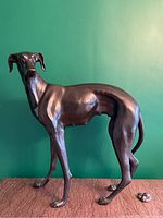 Front three-quarter view of life-size bronze greyhound statue on wooden surface against green background.