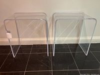 Two clear lucite end tables side by side against wall on black tile floor