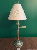Full view of the silver toned metal table lamp with conical, floral cut paper shade on a brown surface and green background.