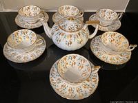 Overall view of six teacups with saucers arranged around central teapot on black surface.