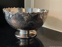 Full exterior front view showing scalloped rim, grape vine relief border, engraved scrollwork, and pedestal base