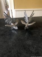 Pair of silver-tone metal rooster figurines in fighting pose shown from front angled view on dark surface.