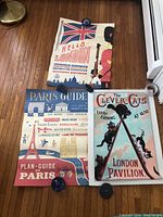 Three paper travel posters laid on hardwood floor, one showing a British Guard with Union Jack flag saying Hello London, another a Paris travel guide map, and the third a vintage cat show poster titled The Clever Cats.
