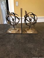 Pair of brass sundial armillary sphere bookends side by side, showing front view, polished brass with circular rings and central arrow design.