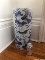 Full view of the tall cylindrical porcelain umbrella stand next to a soda can for scale, showing blue floral and bird pattern on white background.