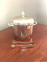 Photo showing full stainless steel ice bucket with lid and attached decorative handles, and ice tongs in front on wooden surface.