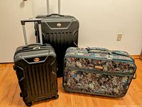 Three suitcases displayed on hardwood floor against white wall: two black hard cases and one fabric tapestry suitcase.