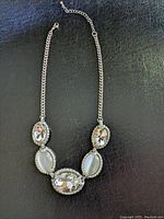 Full view of silver tone chain necklace featuring five oval stones, set with rhinestone edging, flat laid on black surface.