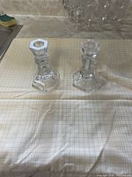 Pair of clear crystal candle holders on grid-pattern cloth background, front view showing hexagonal base and body details.