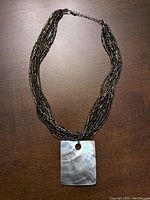 Full view of the multi-strand glass seed bead necklace with attached rectangular Mother-of-Pearl pendant on a wood surface.
