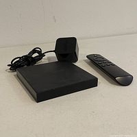 Front view of Amazon Fire TV device, power adapter, and remote on white surface.