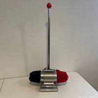 Full view of the Dremel shoe buffer polisher showing vertical rod with red handle and dual rotating brushes in black and red on metal base.