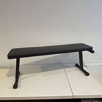 Side view of folding weight bench showing rectangular padded black seat and metal tubular legs with rubber caps.