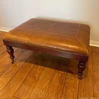 Front angle view of leather ottoman showing wooden legs with brass caster wheels and decorative leather stitching.