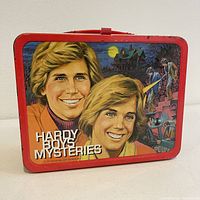 Front side of the Hardy Boys Mysteries lunchbox showing two characters with a haunted house scene in the background, red metal frame and plastic handle on top.