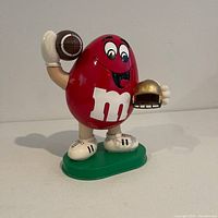 Front view showing the Red Peanut M&M as a football player holding a football and a gold helmet in hand, standing on a green base.