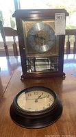 Photo showing both clocks together on a table: Ansonia mantel clock with glass front case and separate black wall clock face laid flat.