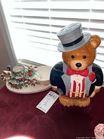 Front view of bear-shaped cookie jar in tuxedo and Christmas-themed platter with cup and pitcher on a table near window