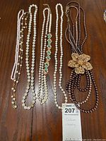 Photo showing seven costume jewelry necklaces on a wooden surface, featuring several pearl-like strands, a crystal chain, and a dark multi-strand beaded necklace with floral clasp.