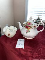 Photo of white ruffled hand painted Fenton dish with cherry design next to a Royal Albert Old Country Roses teapot with rose motifs and gold trim.