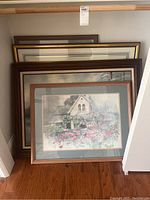 Five framed wall art pieces stacked against a wall with visible floral cottage painting and adjacent abstract landscape artwork