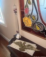 Galileo thermometer standing on wooden furniture with brass eagle wall art lying flat on a cloth nearby. Wall stained glass art in background.