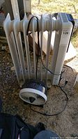 Full view of the white radiator oil heater and the black and white fan placed on the ground outdoors