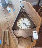 Front and angled shots showing the full cuckoo clock including the clock face, wooden pendulum panel, weights, and chain.