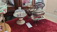 Front angle of the full silver tea set on red cloth including a pedestal tray with glass dome, pitcher, and teapot on burner stand with black handle.