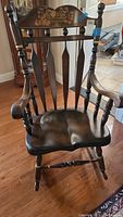 Front view of a dark wooden rocking chair with carved floral design on top backrest showing full seat, armrests, and rocker base.