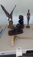 Four statues including eagle, thinker figure, abstract human figure, and dog figurine flat on table with lot tag visible.