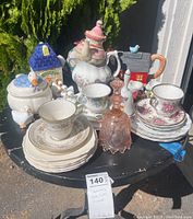 Photo showing all items on a round black table with trees in background. Items include teapots, tea cups on saucers, glass bell, decorative houses and white bird figurines.