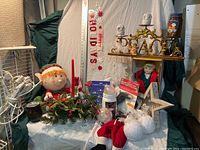 Full view of Christmas decorations lot including elf doll, ceramic doll, candle holder with red taper candle, Santa pants bottle cover, card magnets, ladder lights on tester, and 'PEACE' ornament.