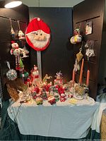 Wide view of table displaying numerous Christmas decorations including the large Santa face wall hanging, small Santa and snowman figurines, candle holders, and other holiday items on a white cloth.