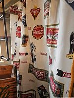 Fabric shower curtain hanging on a rod featuring multiple Coca-Cola logos and vintage graphics in red, green, black and cream.