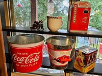 Wide shot showing two metal Coca-Cola ice buckets, two rectangular Coca-Cola tin cans, a white ceramic mug with checkered rim, and Coca-Cola bottle-shaped shower curtain rings