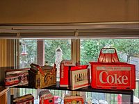 Three Coca-Cola collectible tins in various shapes with vintage designs displayed on a shelf by a window along with a deck of Coca-Cola playing cards and the red plastic crate caddy visible to the right.