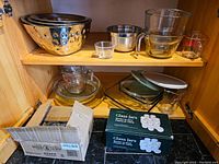 Shelved kitchenware showing nested metal and glass mixing bowls, glass baking dishes, a set of measuring cups, and unopened boxes of glass spice jars.